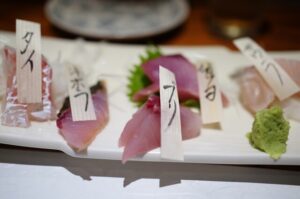 地下鉄赤坂駅から徒歩約3分。福岡市中央区舞鶴の「酒場・食事処 魚の まる田（まるた）」は、地元の食通たちからも厚い支持を集める魚介料理の専門店です。店主自ら市場で仕入れる魚は、季節や天候によってラインナップが日々変わり、訪れるたびに違った旬の味覚を楽しめるのが魅力です。 九州の旬魚が主役のおまかせ刺身コース 看板ともいえるのが、おまかせコースで提供される刺身の盛り合わせ。対馬や五島、玄界灘など九州近海で水揚げされた魚を中心に、店主がその日最も状態のよい魚を厳選。盛りつけには寿司職人としての経験が活かされており、厚みのある切り身は食感と旨みのバランスに優れています。さらに、煮つけや焼き物、茶碗蒸し、小鉢などが順に供される構成で、コースの最後には握り寿司が登場！ネタに合わせて酢飯の温度や握り加減まで調整されており、細やかな技術が光ります。 料理に寄り添う辛口中心の酒と飲み放題プラン お酒はビールや焼酎のほか、日本酒も数種用意されており、料理に合うよう辛口を中心にセレクトされています。また、グループで訪れる場合には、飲み放題付きのおまかせコース（2時間制）もあり、料理内容に応じて柔軟に対応してくれるのも嬉しいポイントです◎ 木の温もりと職人技を間近に感じるくつろぎ空間 店内はカウンター席とテーブル席を中心に全19席。落ち着いた照明と木の温もりを感じる設えで、初めてでもくつろげる雰囲気です。奥には仕切り付きの半個室スペースもあり、4～8名程度のグループ利用にも適しています。料理人との距離が近いカウンター席では、手際よく魚を捌く様子を眺めながら食事ができ、料理の一皿一皿に対する丁寧な所作に引き込まれる時間が流れます！
